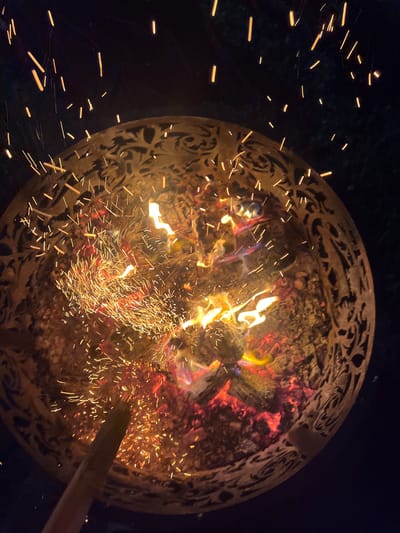 A fire burns in a decorative metal fire pit at night. Sparks fly upwards from the fire, against a dark background. The fire pit has an intricate, carved design around its rim. A wooden stick is visible in the lower left of the image, appearing to be used to tend the fire. The scene is likely an outdoor setting, possibly a backyard or patio. The city of Popovka, Russia is not depicted in the image.