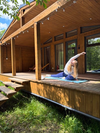 A woman with long blonde hair, wearing sunglasses, a white long-sleeved top, and blue yoga pants, is performing a yoga pose on a blue mat. She is on a wooden deck next to a modern wooden house, with her right arm extended upwards and her left hand near her left foot, executing a stretching posture akin to Extended Side Angle Pose.

The setting is outdoors on a sunny day, with strong shadows cast by the wooden structures, suggesting either morning or late afternoon. The house is constructed from warm-toned wooden planks and features numerous large windows with dark frames, reflecting the surrounding green trees and the blue sky. String lights with circular bulbs are strung along the overhead wooden beams of the deck, which also has a wooden staircase leading down to a natural grassy area with lush green vegetation. Inside the house, visible through one of the windows, is a wooden chair. The number "17" is visible on the wooden frame above a window on the house's exterior. The scene portrays a calm moment of exercise in a tranquil, nature-integrated environment.