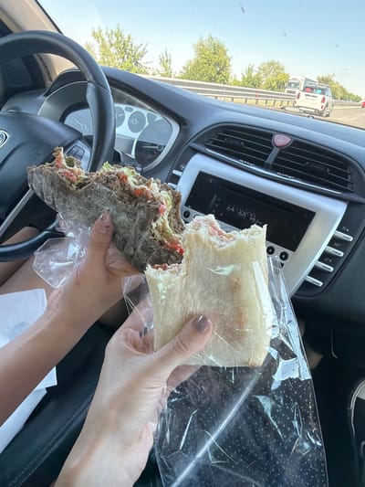 The image shows a close-up view of two hands holding parts of a large sandwich inside a car. The car's dashboard, steering wheel, and radio are visible in the background. The sandwich appears to be made with dark bread and filled with lettuce, tomatoes, and other ingredients. One hand is holding the larger, intact portion of the sandwich, while the other hand holds a bitten-off piece, both wrapped in clear plastic. The scene suggests the individuals are on a road trip, possibly passing through or near the city of Voyvodino, Bulgaria, as indicated by the road and surrounding vegetation. The sunlight suggests it is daytime.