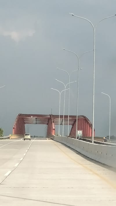 The image captures a view from a vehicle traveling on a highway in Gohor Lama, Indonesia. A distinctive red arched bridge dominates the background, with a single white car visible approaching it on the highway. Numerous streetlights line the right side of the highway, extending towards the bridge. A speed limit sign indicating "40" is positioned on the right side of the road near the bridge. The sky above is overcast, contributing to a muted, grey atmosphere. The highway itself is paved and marked with white lane dividers. The scene appears to be an exterior shot taken during daylight hours.