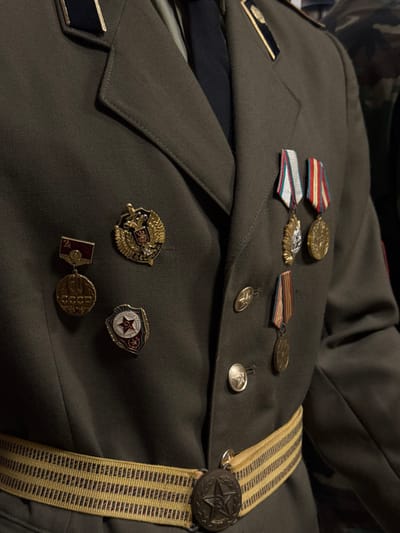 A green military uniform is adorned with various medals and insignia. Ribbons with attached medallions are pinned to the upper chest. Smaller, decorative pins featuring eagles, stars, and Soviet symbols are clustered below. A striped, tan-colored strap with a large, star-shaped button runs vertically along the fabric. Dark shoulder boards are visible, featuring gold accents.