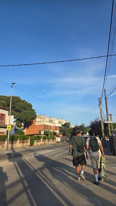 The image depicts a street scene in Santa Perpètua de Mogoda, Spain, on a bright, clear day, likely late afternoon given the long shadows and visible daytime moon. The composition is oriented vertically, showing a wide expanse of blue sky, a paved road, and buildings lining one side.
Two young men are positioned on the asphalt road, possibly on a faded crosswalk. The man in the foreground is dressed in a dark green t-shirt, dark shorts, white socks, and white sneakers with green accents, and wears a dark crossbody bag. A tattoo is visible on his left arm. He holds a skateboard. Behind him, another man wears a light-colored t-shirt, light shorts, white socks, and dark sneakers, carrying a dark backpack with a water bottle. He is also holding a skateboard, the underside of which features a green graphic. They appear to be walking or standing, not actively riding their skateboards.
The right side of the road is bordered by multi-story residential buildings with light-colored facades and some red brick details, interspersed with green hedges and trees. Overhead utility poles and numerous power lines traverse the blue sky. A street lamp pole stands on the left side of the road.
The sky is a clear, vibrant blue with a few subtle white clouds. A distinct crescent moon is visible in the upper left portion of the sky. Long, sharp shadows from the people, poles, and unseen objects stretch across the road surface, indicating the sun is low in the sky. Visible text includes the number "32" on a sign attached to a structure in the background and a yellow directional sign with a black arrow pointing right near the street lamp.