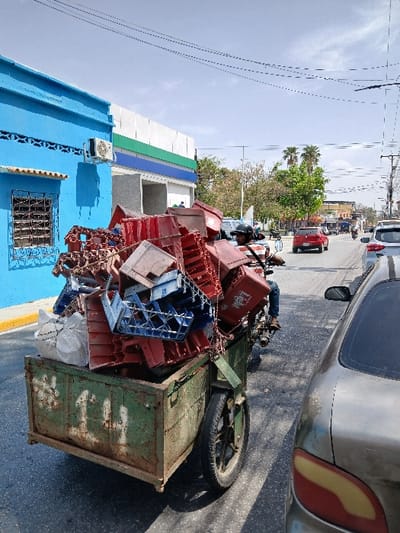 The image depicts a daytime street scene in San Joaquín, Venezuela. In the foreground, a male individual, wearing a dark helmet, dark shirt, and blue jeans, operates a motorcycle towing a two-wheeled, rusted green metal trailer. The trailer is heavily loaded with numerous red and several blue plastic crates, along with a white sack, piled high above its sides. Dark straps or chains appear to secure the load. To the immediate right, the rear quarter of a silver/grey passenger vehicle is visible, featuring a combined red and amber taillight cluster. On the left side of the street, a bright blue building with an externally mounted white air conditioning unit and a barred window stands adjacent to a commercial structure characterized by a horizontal white, green, and blue striped facade. A yellow painted curb is visible along the left side of the roadway. The asphalt road extends into the middle distance, showing several other vehicles, including a red car. Overhead utility lines are present against a mostly clear blue sky. Palm trees and other foliage line the street further in the background.