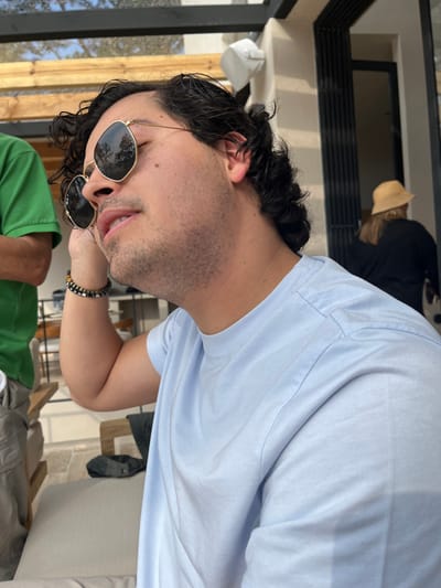 A man with curly black hair and sunglasses tilts his head, facing the camera with a slight smile. He's wearing a light blue t-shirt and a beaded bracelet. The background shows glimpses of an outdoor setting, including a wooden structure, another person in a straw hat, and someone wearing a green shirt. The picture appears to be taken from a slightly off-kilter angle indoors or on a patio.