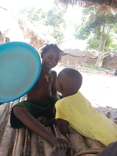 Two young children are seated on a makeshift wooden bench in what appears to be a rural village setting. The child on the left, facing partially towards the viewer, holds a large, light blue plastic basin. They are wearing a dark green garment and a gold-colored cross necklace. Their hair is styled in braids. The child on the right is facing away from the viewer, wearing a bright yellow shirt. Both children appear to have dark skin and short, dark hair.
The background suggests a simple, possibly impoverished, environment with structures made of thatched roofs and natural materials. Trees and foliage are visible, indicating an outdoor setting. The lighting suggests it is daytime, with bright sunlight filtering through the trees, creating some overexposed areas in the image. The ground appears to be dirt or sand, with some scattered debris. The overall scene conveys a sense of everyday life in a community with limited resources. No visible text is present in the image.