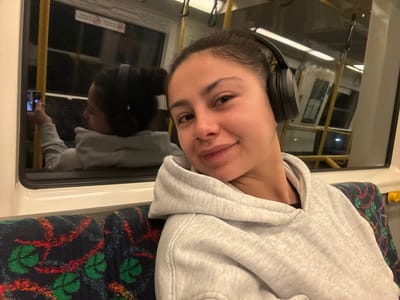 The image shows a young woman sitting on a public transport vehicle, likely a train, in Bayswater, Australia. She is wearing a grey hooded sweatshirt and black headphones, and she is holding a smartphone in her left hand, seemingly taking a selfie. The train's interior is visible, with patterned seats and windows reflecting the outside environment. The lighting suggests it is either evening or a dark day. The woman has a nose ring and her hair is pulled back. She is smiling at the camera.