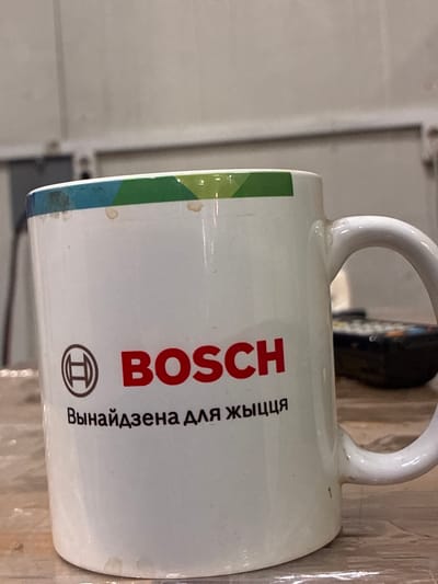 A white ceramic mug with the Bosch logo and text is placed on a wooden surface. The mug has a colored geometric pattern around its rim, featuring blue, green, and yellow segments. The Bosch logo, consisting of a circular symbol and the word "BOSCH" in red, is prominently displayed on the mug. Below the logo, text in Cyrillic script reads "Вынайдзена для жыцця" (Vynaydzenа dlya zhytstsya), which translates to "Invented for life". The mug appears to have some minor smudges and stains on its surface and rim. In the background, out of focus, a remote control and a wall are visible. The setting suggests an indoor environment, possibly a home or office. The scene does not provide any specific indicators of its location within the city of Заслаўе, Belarus, beyond the text on the mug which is the company's slogan.