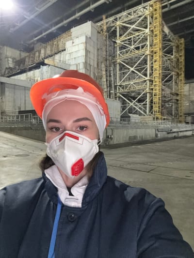 A female individual is depicted in a close-up selfie, centrally framed. She wears an orange hard hat positioned over a white head covering, a white FFP2/N95-type respirator mask with a red exhalation valve covering her nose and mouth, and a dark blue jacket over a white collared shirt. Her eyes are visible above the mask.

The background reveals a large, industrial environment, specifically the Chernobyl Nuclear Power Plant complex, located near the city of Pryp'yat', Kyiv Oblast, Ukraine. To the left, a large, multi-story concrete structure, consistent with a reactor or turbine hall building, is visible. To the right, a tall, intricate metal truss structure, appearing as part of a large scaffolding system or a containment arch like the New Safe Confinement, dominates the upper right background. The area is dimly lit by artificial light, illuminating the subject more brightly than the background. The ground is a flat concrete surface.