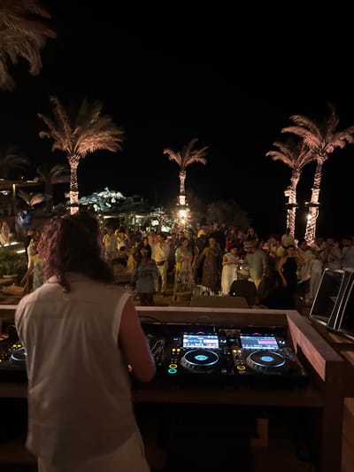 This nighttime image captures an outdoor party in Psarou, Greece. In the foreground, a DJ with long, curly hair, dressed in a light-colored sleeveless shirt and trousers, operates a professional DJ setup. The setup, resting on a wooden stand, includes two Pioneer CDJ-style media players with illuminated jog wheels and screens, flanking a central mixer. Speaker monitors are partially visible to the right.
The midground and background are occupied by a large crowd of individuals, approximately 50-70, socializing and dancing. They wear varied attire, and some hold beverages. The scene is illuminated by warm lights wrapped around multiple palm trees, which stand against a dark night sky. Additional dimly lit structures, likely part of a venue, are visible in the background.