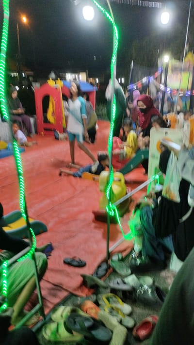 The image depicts an outdoor night scene in Sukodono, Indonesia, possibly at a local event or fair. In the foreground, a collection of various footwear, including flip-flops and sandals, are scattered on a red mat. The background is a blur of activity and lights. A child is lying on their stomach on the red mat, with a green slide and yellow toy structure nearby. Several people are visible, some wearing headscarves. Green string lights are prominent, framing the scene. A red and yellow play structure is partially visible on the left. The overall impression is a lively, somewhat chaotic, community gathering.