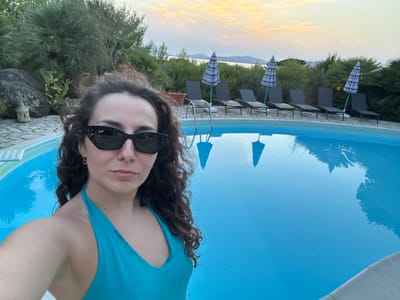 A medium shot captures a woman in a turquoise swimsuit posing for a selfie by a swimming pool at dusk. She is positioned on the left side of the frame, looking directly at the camera with sunglasses on. The pool, filled with bright blue water, occupies the majority of the right side of the image. Beyond the pool, a row of lounge chairs and closed umbrellas are set up, backed by dense green vegetation and trees. In the distance, a body of water is visible, with hills or mountains on the horizon under a pale orange and yellow sky. The overall scene suggests a serene outdoor setting, likely at a resort or private residence, possibly in the Alghero area given the coastal landscape and Mediterranean flora.
