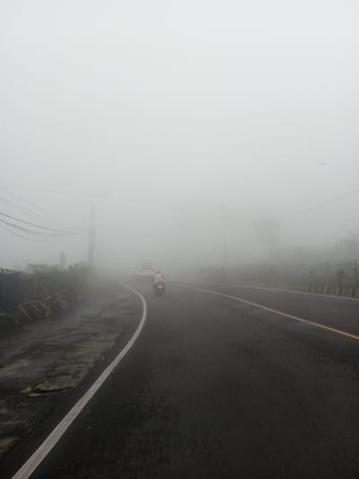 A road disappears into dense fog, obscuring visibility. A white car and a single motorcyclist venture into the misty distance. The road is asphalt with yellow and white lane markings. To the left, a concrete barrier and overgrown vegetation line the road's edge. On the right, a fence and some trees are barely visible through the fog. Overhead wires stretch across the sky. The pervasive fog creates a sense of mystery and limited perspective.