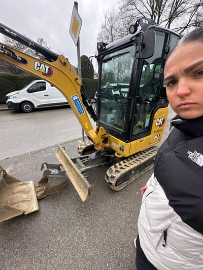 This image depicts a street scene in Grünwald, Germany, under an overcast sky. A female subject, positioned on the right side of the frame, is taking a selfie, looking directly at the camera with a neutral expression. She is wearing a black and white "The North Face" puffer jacket. To her left, a yellow Caterpillar 301.8 compact excavator, prominently branded with "CAT," "Zepellin," and "PFAFFINGER" decals, is stationary on the asphalt roadway. Its tracks, dozer blade, and attached digging bucket show evidence of dirt and mud residue. Behind the excavator's boom, a yellow diamond-shaped priority road sign on a grey pole is visible. In the background, a white van is parked or moving on the street, bordered by a green hedge and bare trees. The asphalt surface appears wet and contains scattered debris.