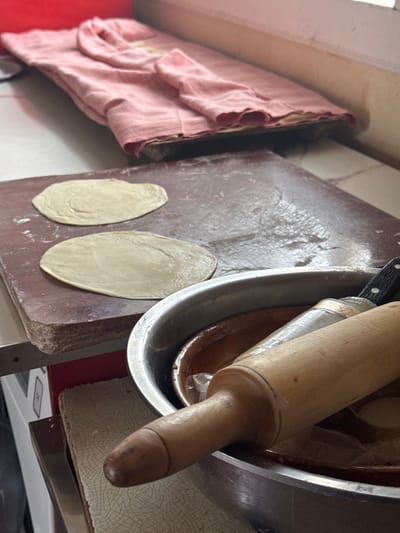 This image depicts a food preparation scene in Dehqonbozor, Uzbekistan. In the midground, two flattened, light-colored circular pieces of dough are placed on a dark reddish-brown wooden cutting board, which exhibits a visible sheen suggesting oil or moisture. In the foreground to the right, a large metallic bowl contains a light-colored wooden rolling pin and a knife with a black handle. A reddish-brown ceramic bowl is partially visible within the metallic bowl, behind the rolling pin, with some translucent plastic material visible around it. The background features a folded pink garment resting on an elevated wooden shelf or surface, positioned against a plain light-colored wall. No individuals are present in the frame.