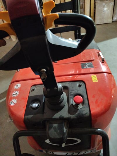 This is a close-up, ground-level, eye-level, exterior, medium shot of the control panel and handle of an orange electric pallet jack. The pallet jack is indoors, in what appears to be a warehouse or storage facility. The environment is dimly lit, with a concrete floor and shelving units visible in the background. There are several warning stickers on the pallet jack, including a pictogram of a person falling, a pictogram of a person carrying a load, and a pictogram of a person walking. There is also a sticker with an exclamation mark and an open book, indicating caution and a manual. A red emergency stop button is visible on the control panel. The pallet jack has a black steering handle with yellow grips. The overall condition of the pallet jack appears to be used, with some dust and scuffs visible on its surface. There are no people or animals visible in the image. The pallet jack is not actively being used; it is stationary. The image does not provide any information about the location within the city of Okhtyrka, Ukraine.