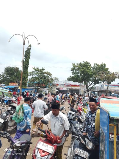 A crowded street scene features people wearing traditional Muslim attire, with many sporting black peci hats. A cluster of parked motorbikes fills the foreground, showing license plates. A tall lamp post wrapped in vines stands to the left. In the background, an “Alfamart” convenience store is visible. The image, captured with a Vivo Y19s, includes a timestamp of February 6, 2026.
