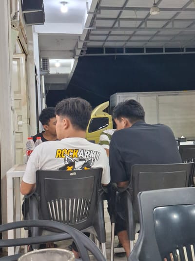 In Sigli, Indonesia, three individuals are seated at tables outdoors, under a covered area with fluorescent lighting. Two men are facing away from the camera, seated at a table in the foreground. The man on the left wears a white t-shirt with the text "ROCKARMY" printed on the back in yellow and black, alongside a graphic of a cat or wolf-like creature. He is seated in a black plastic chair with a slatted back. To his right, another man wears a black shirt and is also seated in a black plastic chair. A third individual, partially visible to the left, faces the other two and is wearing a black t-shirt. Two plastic water bottles are visible on the table in front of the man in the white shirt. In the background, a yellow truck and a white cargo truck are parked. The environment appears to be a food stall or restaurant area.