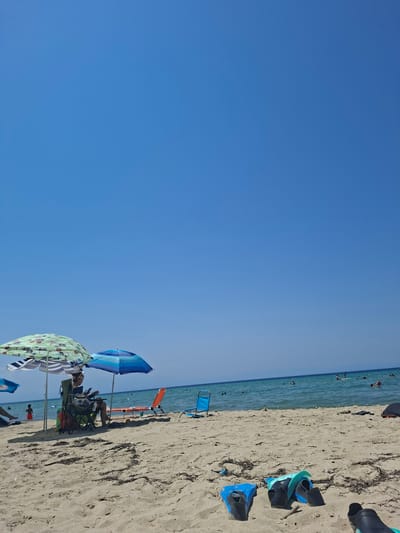 The image depicts a sunny day on a sandy beach in Nea Kallikrateia, Greece. The sky is a clear, vibrant blue. In the foreground, several pairs of flippers are laid out on the sand.

Further back on the beach, two beach umbrellas provide shade. Beneath one umbrella, a person is seated in a folding chair, appearing to read. Next to them, an orange beach chair is empty. A few other beach chairs and umbrellas are visible further down the beach.

The sea is a calm, turquoise color, with several people swimming and engaging in water activities. The shoreline is a mix of sand and some dried seaweed. The overall scene conveys a relaxed atmosphere typical of a summer day at the beach.