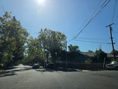 This media file depicts a residential street scene in Sacramento, California, under a bright, sunny sky. The time of day appears to be late morning or early afternoon, with the sun high and casting distinct shadows. The weather is clear and warm, indicated by the bright sunlight and the presence of lush green trees lining the street.
The scene is dominated by a two-lane asphalt road with several cars parked along the sides and a few in motion. The architecture of the houses visible suggests a suburban setting, with some single-story homes and a mix of architectural styles. A wooden fence runs along the property line in front of one of the houses. Overhead, power lines stretch across the sky, supported by a wooden utility pole.
One notable detail is a street sign at an intersection, clearly visible and reading "Santa Cruz Ave". The presence of mature trees with dense foliage suggests that this is a well-established neighborhood. The overall mood is peaceful and ordinary, capturing a typical day in a residential area. There are no people visible in the frame.