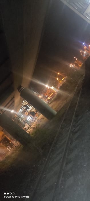 The image presents a nighttime outdoor scene in the city of Constance, Germany, taken from a tilted perspective approximately 45 degrees clockwise. In the central foreground, a light-colored cylindrical object, resembling a thermos or large bottle with a dark cap, rests horizontally on rough ground. Adjacent to this object and extending into the lower right quadrant are two parallel railway tracks. To the left, a large, dark concrete structural element, possibly part of a bridge or underpass, occupies a significant portion of the frame. The background features various distant artificial light sources from buildings and streetlights, indicating an urban or industrial area beyond the immediate railway environment. Sparse, dark vegetation is visible between the foreground and the illuminated background. No individuals or specific actions are depicted.