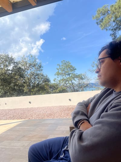 A person with glasses looks down, wearing a gray sweatshirt and blue pants. They are standing next to a light-colored wall with a textured section. The background shows a blue sky with white clouds and green trees. Part of a building's roof is visible in the lower left corner. The bracelets are visible on the left wrist of the person.