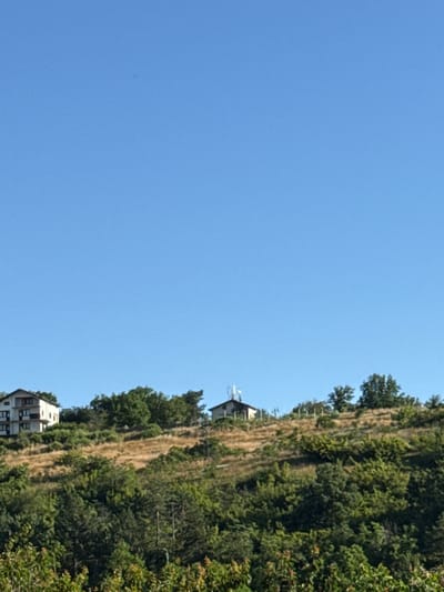 The image shows a rural landscape with two houses on a hillside under a clear blue sky. The house on the left is larger and appears to be a multi-story building with several windows and balconies. The house on the right is smaller and has a communication tower on its roof. The hillside is covered with a mix of dry grass, green bushes, and trees. The scene is likely located in the vicinity of Slavyani, Bulgaria, given the context.