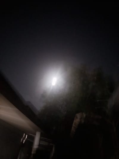 The image depicts a night scene with a bright, elongated light source in the upper center, possibly a streetlamp or a distant light, casting a glow through a hazy atmosphere. Silhouetted against this light are dark shapes of trees and foliage. In the lower left, a blurred diagonal structure, likely a building or a roofline, is visible, also rendered indistinct by the low light and motion blur. The overall impression is one of an outdoor urban or semi-urban environment at night, with the lighting and atmospheric conditions creating a somewhat abstract and moody effect. The location in Tibojong, Indonesia is not discernible from the visual information provided in the image itself.