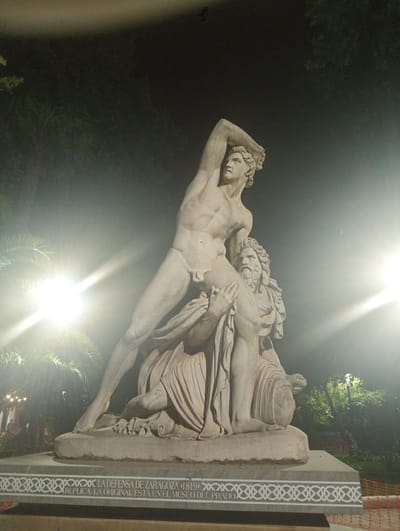 The image shows a nighttime view of a large, light-colored marble statue depicting two male figures. The statue is illuminated by bright, possibly artificial, lights, creating lens flares and bright spots in the scene. The figures are in an active pose; one figure stands over another who appears to be kneeling or seated, with the standing figure raising an arm as if to strike. The statue is mounted on a rectangular base with decorative white patterns along its sides. Text is visible on the base, reading "LA DEFENSA DE ZARAGOZA *1819* RÉPLICA. LA ORIGINAL ESTÁ EN EL MUSEO DE". The statue is situated outdoors, with dark trees and foliage in the background, some of which appear to be palm trees. There is also some orange construction fencing visible in the lower right corner of the image. The scene is located in Priego de Córdoba, Spain.