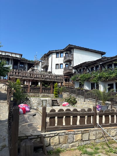 This image depicts a sunny daytime scene in Medovo, Bulgaria, showcasing a cluster of buildings in a traditional architectural style. The multi-story structures feature white plastered walls and prominent dark wooden elements, including ornate balconies, exposed beams, and railings. Lush green vines are draped across some of the upper balconies, adding a natural, vibrant touch. Numerous windows, some reflecting the blue sky, are visible across the facades.
The ground level consists of irregular stone paving, stone steps, and low stone walls, with patches of green grass growing between the stones. Dark wooden fences and balustrades line pathways and elevated areas. Small palm trees and various green plants, including bushes with delicate pink flowers, are interspersed throughout the landscape.
The sky is a clear, bright blue, indicating fair weather and strong sunlight. Distinct shadows are cast across the buildings and ground, suggesting either a mid-morning or late afternoon time of day. Two round, patterned objects, primarily red and white, resembling cushions or poufs, are resting on the stone ground near the bottom right of the frame, alongside a purple and white bag. A white pipe and some hoses are visible near a set of stone steps. The scene appears calm and peaceful, with no people visible. No discernible text is present in the image.