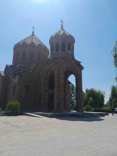 The photograph shows the Saint Gregory Cathedral in Artashat, Armenia. The cathedral is a large, stone building with two domed towers, the larger of which is topped with a cross. The building is made of reddish-brown stone and features arched doorways and windows. In the foreground, there is a paved courtyard with some green bushes and trees. The sky is clear and blue. In the background, some people can be seen walking in the courtyard. The overall impression is of a sunny day at a historic religious site.