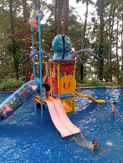 An outdoor water play area is depicted in Kuwukan, Indonesia. The scene features a multi-tiered, colorful water park structure set within a blue swimming pool. The structure includes yellow, red, and blue platforms, with a large teal and blue octopus figure atop. A tall pole supports a large, multicolored tipping bucket. Three slides descend into the pool: a large blue slide adorned with cartoon characters on the left, a pink double-lane slide in the center, and a yellow curved slide on the right. One individual, dressed in dark attire, stands on a platform of the structure near the pink slide. In the pool, one child is partially submerged, swimming in the foreground, while a second child stands in the shallower water in the background. Tall evergreen trees form a dense forest behind the water park, indicating a natural or wooded environment.