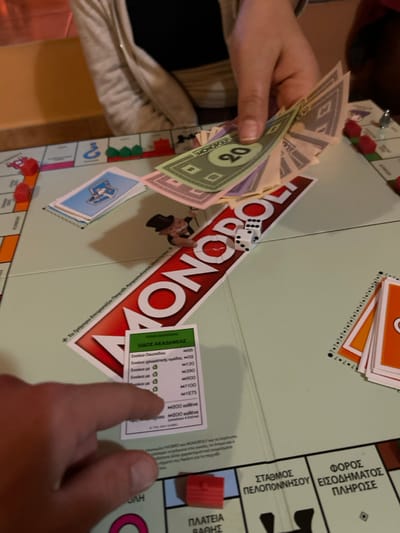 An overhead, close-up view depicts a Monopoly board game in progress, illuminated by indoor lighting. A fair-skinned hand with visible fingernail holds a fan of Monopoly currency, prominently displaying a green 20-unit bill. Directly below, two white dice rest on the red "MONOPOLY" logo, showing faces 3 and 4. Another fair-skinned hand, with a visible thumb and index finger, points to a "ΟΔΟΣ ΑΚΑΔΗΜΙΑΣ" (Akadimias Street) property card, which details rent information in Greek.
The light green game board features various Greek-labeled spaces including "ΣΤΑΘΜΟΣ ΠΕΛΟΠΟΝΝΗΣΟΥ" (Peloponnese Station), "ΦΟΡΟΣ ΕΙΣΟΔΗΜΑΤΟΣ ΠΛΗΡΩΣΗΣ" (Income Tax Payment), and a blue "Chance" card pile. Scattered on the board are several red plastic houses/hotels and three green plastic houses. A stack of orange cards lies on the right side of the board. The blurred background shows a beige or light brown surface, likely a table, and the upper body of a person wearing a light-colored cardigan. The scene is located in the city of Gerakas Municipal Unit, Greece, as indicated by the Greek language Monopoly board and the specified location.