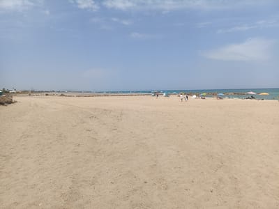 This image depicts a wide, sandy beach on a clear, sunny day. In the foreground, the sand is mostly empty, showing some tire tracks and footprints. In the middle ground, there are several people relaxing on the beach, with many colorful umbrellas providing shade. Some individuals are walking along the shore, while others are seated or lying down. The water is calm and blue, meeting the sand at a distant breakwater. The background shows a hazy coastline with some buildings and vegetation, suggesting a populated area. The overall atmosphere is one of a typical summer day at the beach, with people enjoying leisure time. The time of day appears to be afternoon, given the bright sunlight and the shadows. The weather is warm and pleasant, with a few wispy clouds in the pale blue sky. The location is likely Sagunto, Spain, as indicated by the context. There is no visible text in the image.
