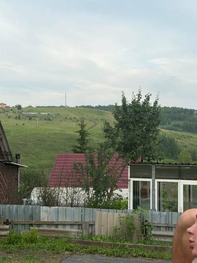 The image depicts a rural landscape with a gently rolling green hill in the background, dotted with trees and a winding dirt path. Several houses and buildings are visible on the hillside, along with a tall transmission tower. In the foreground, there is a weathered corrugated metal fence, with a wooden structure and green vegetation. A red-tiled roof of a building is partially visible to the left of center. A greenhouse or covered structure with glass panels is situated in the mid-ground. The sky is overcast with a pale blue hue. A portion of a person's shoulder and cheek is visible in the lower right corner of the frame. The scene is consistent with a view from a residential area in Кузнецово, Russia, likely a dacha or private property, given the rural setting and presence of residential structures.
