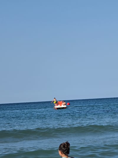 The image depicts a serene scene at sea, likely near Ahtopol, Bulgaria, on a clear, sunny day. The deep blue expanse of the sea meets a pale blue sky, creating a vast horizon. In the middle distance, a small white and orange pedal boat is visible, occupied by at least two people. One individual, wearing yellow shorts, is in the process of jumping from the boat into the water, captured mid-air. Another person, possibly a child in a pink bathing suit, is seated on the boat. A red buoy floats nearby. In the foreground, the back of a person's head, with their hair tied up, is visible, as they are partially submerged in the water, facing away from the camera. Gentle waves create a textured surface on the sea.