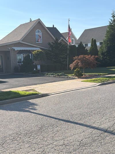 The image shows a residential street in Levittown, New York. A two-story house with brown siding and a gray roof is visible on the left side of the frame. An American flag flies from a flagpole in the yard in front of the house, with a smaller red flag beneath it. The house has an arched window on the upper floor with an American flag decoration visible inside. In front of the house, there is a neatly manicured lawn with various shrubs and a small decorative tree. A sidewalk runs along the street, separating the road from the properties. The road is paved with asphalt and shows some cracking. The sky is clear and blue, indicating it is a sunny day. The overall scene depicts a well-maintained suburban environment.