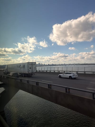 The image depicts a view from the Samuel De Champlain Bridge in Brossard, Canada. The bridge spans the Saint Lawrence River. The sky is visible with scattered clouds. A white car is traveling on the bridge. A long Vantage Logistics truck is also present on the bridge. Below the bridge is the river water. The cityscape can be seen in the distance.