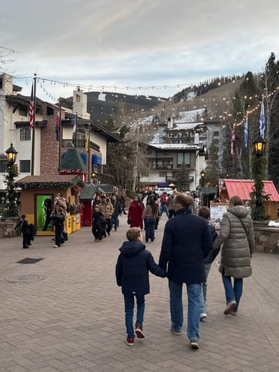 A wintry street scene features a crowd of people walking on a brick-paved path. Colorful buildings line the left side, adorned with holiday decorations, flags, and lights. A ski slope with sparse snow cover rises in the background, also decorated with lights. A family is walking towards the camera, including a child holding the hand of an adult. Overcast sky suggests a cold day.