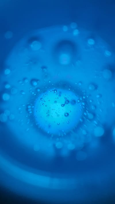 This is a close-up, abstract photograph of water with many small bubbles and larger, blurred orbs, all rendered in shades of blue. The overall effect is reminiscent of looking through a liquid at something illuminated from within or behind. There are no discernible people, objects, or a clear scene or setting. The image is abstract, focusing on the interplay of light and liquid. The activity taking place is the formation or presence of bubbles within a fluid.
Notable details include the varying sizes of the bubbles, some appearing as sharp circles and others as soft, diffused lights. There's a central area of brighter illumination, possibly a light source or a larger bubble, surrounded by smaller ones. The background is a deep, solid blue, suggesting a controlled environment or a deep body of water, though no specific location cues are evident. There is no indication of time of day or weather. The emotion evoked is one of calm, mystery, or perhaps scientific observation. No text is visible in the image.