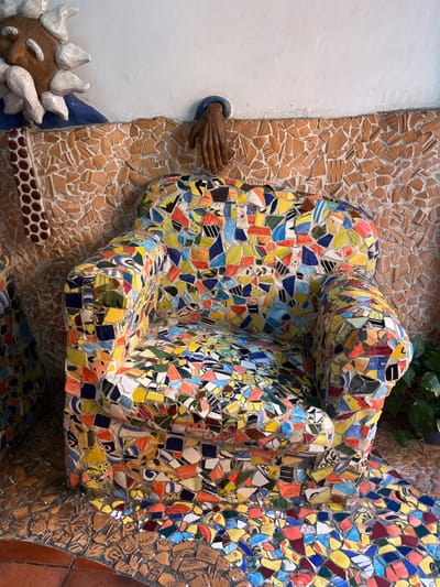 A tiled human-like figure, composed of irregularly shaped colorful pieces, is the focal point. This form is textured with various colors, including yellow, blue, red, and orange. The figure is set against a background made of broken ceramic tiles. A sculpted brown hand reaches out from a white wall, situated to the left of the tiled figure. Underneath, there is a head with white spikes. A touch of greenery is visible in the top right corner of the frame.