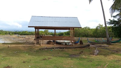 A modest, open-sided wooden structure with a corrugated metal roof stands near a body of water. The structure has a wooden bench running along its interior. A wooden ladder provides access to the raised platform. Several traditional fishing boats, painted in various colors including blue, green, and red, are positioned along the water's edge and on the shore. Some boats are in the water, while others are pulled up on the muddy bank. Bags, possibly containing fishing gear or supplies, are visible near the boats. The environment is a coastal area with lush green trees in the background, suggesting a tropical setting. A palm tree stands to the right of the structure. The ground is covered with grass, dirt, and debris, including fallen palm fronds and scattered litter. The sky is overcast with some clouds. The scene depicts a typical fishing village environment in Pusong, Indonesia, characterized by its traditional boats and waterfront structures.