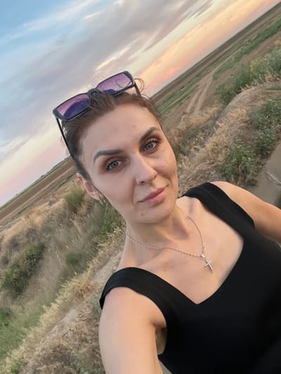 A medium shot, outdoor selfie captures a woman with light skin, medium brown hair styled up, and blue eyes looking directly at the camera. She is wearing a black sleeveless top, a silver necklace with a cross pendant, and sunglasses perched on her head. Her makeup includes defined eyebrows, eyeshadow, and lipstick. The background shows a dry, grassy landscape with fields and a muted sky with hints of pink and blue, indicative of dusk or dawn. The location is identified as Aqsu city administration, Kazakhstan.