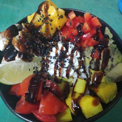 This is a close-up, overhead shot of a colorful poke bowl presented in a black bowl. The bowl is filled with a variety of ingredients, including diced red bell pepper, cubed mango, and sliced avocado. A dollop of white sauce, possibly sour cream or mayonnaise, sits in the center, drizzled with a dark sauce, likely soy sauce or a teriyaki glaze. Black sesame seeds are sprinkled generously over the entire dish. There are also chunks of what appears to be grilled chicken or fish underneath some of the vegetables, also coated in the dark sauce. A slice of lime is visible on the right side of the bowl. The food is arranged in a visually appealing, segmented pattern within the bowl. The background is a teal or dark green surface, possibly a countertop or placemat. The lighting suggests an indoor setting. There is no visible text in the image. The context provided indicates the location is Kobeliaky, Ukraine, suggesting this is a dish from a restaurant or prepared in a home kitchen there. The image focuses solely on the food, with no people or other objects prominently featured.
