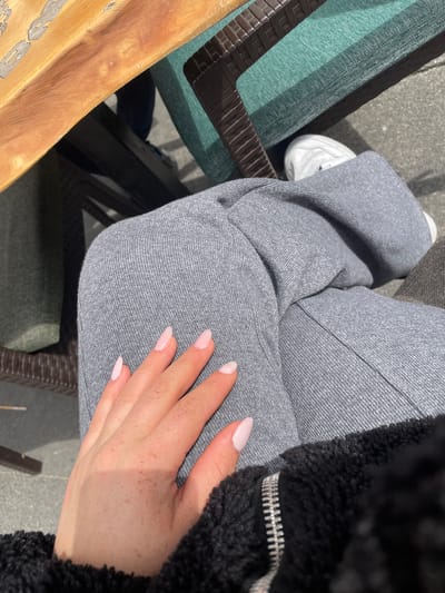 This media file captures a close-up, high-angle view of a person seated outdoors on a bright, sunny day in Dortmund, Germany. The subject is primarily the person's lower body, seen from the waist down, with their left hand resting on their lap.

The person is wearing light grey, possibly heathered, sweatpants or track pants, which show subtle fabric texture and seams. A white sneaker is visible on their left foot. On the right side of the frame, a black, fuzzy or sherpa-style jacket is partially visible, featuring a prominent silver zipper.

The person's left hand, with light pink or nude-colored manicured nails, is casually placed on their lap. The skin on the hand exhibits a scattering of small freckles. Bright sunlight casts a noticeable shadow across the hand, indicating clear, sunny weather, likely during the day.

The setting appears to be an outdoor area, possibly a patio or a cafe. Part of a wooden surface, potentially a table or chair arm, is visible in the bottom left corner. Adjacent to it, a chair with dark brown, possibly woven, structural elements and a cushion upholstered in green and dark brown striped fabric can be seen. The ground is a light grey, textured surface, resembling concrete or pavement. The image conveys a sense of casual relaxation. There is no visible text in the image.