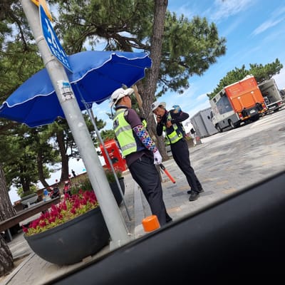 In Gangneung-si, South Korea, two individuals wearing high-visibility vests and white hats are interacting near a pole with a blue umbrella. One individual is facing left, holding an orange signaling stick, and appears to be speaking. The other individual is facing right and speaking into a radio. Behind them, a bright orange truck with a white trailer is parked, and to the left, a red postal box is visible. The ground is paved with a brick pattern. Pine trees and a bright blue sky with some clouds form the background. Red flowers are planted in a large black pot in the foreground.