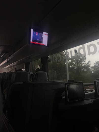 The image is taken from the interior of a bus looking forward, with the overhead compartments and a window visible. The bus is parked in a wooded area in Guadarrama, Spain, at the coordinates provided. The window reveals a heavily overcast sky, with trees on the other side. There are five rows of seats, with the backs of the seats visible. The seats are black and covered in fabric. The bus appears to be empty. A flat-screen monitor is visible on the ceiling of the bus, with a digital screen in the upper right corner. The screen is turned on and displaying a colorful image. The word "apre" is visible on the white ceiling of the bus, possibly a logo or part of a sticker.  The bus appears to be a modern, comfortable one. There are no people present in the scene.