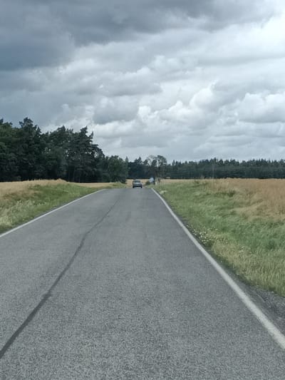A rural road curves through a landscape of fields and trees under a cloudy sky. A single dark-colored car is visible driving away from the camera in the distance on the right side of the road. To the left of the road, a field of dry, yellow grass stretches towards a dense line of dark green trees. The sky is filled with heavy, gray clouds, suggesting overcast or impending rain. The road itself is paved and shows a visible seam running down the center. The scene appears to be a quiet, sparsely populated area, likely in the Czech Republic, possibly near Čistá.