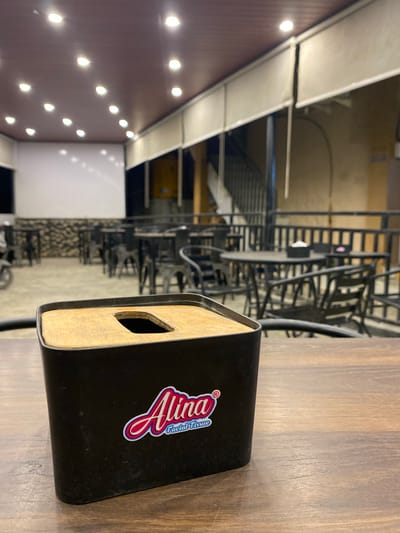 The image captures a scene within a restaurant or eatery in Keudeublang, Indonesia. A tissue box labeled "Alina Facial Tissue" sits prominently on a wooden table. The environment seems to be at a place where they keep their tables and chairs. The background contains other tables and chairs. The lighting in the space consists of recessed ceiling lights.