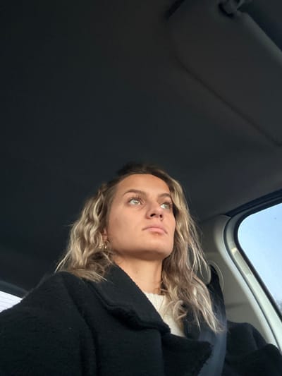 A young woman with wavy blonde hair and green eyes looks up, seemingly from inside a car. She is wearing a black coat over a light-colored top. The car's interior is visible, including the sunroof and the sun visor. The angle of the shot is from below, emphasizing the woman's upward gaze. The background is mostly dark.