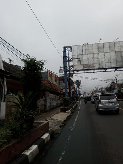 The image displays an overcast daytime street scene. A multi-lane asphalt road occupies the center and right, featuring a faded white dashed line and black-and-white striped curbs. A silver Toyota Avanza (license plate F 1749 NY) is positioned on the right lane, facing away from the viewer, with other vehicles and a motorcycle visible in the traffic stream further down the road.
To the left, a concrete sidewalk runs parallel to the road, bordered by a dark brown planter box containing green foliage, including potted plants. Beyond the sidewalk, a metal fence precedes a series of older, low-rise buildings with red tiled roofs and varied facades, obscured by dense utility wiring in the upper left.
On the right side of the road, a large, unadorned metal billboard structure is mounted on a blue support pole. The billboard is equipped with multiple upward-facing floodlights. Below it, two decorative streetlights with rectangular lanterns are affixed to the same pole. Additional utility poles with numerous power lines and distant buildings line both sides of the street under a uniformly gray sky. No discernible individuals are present.